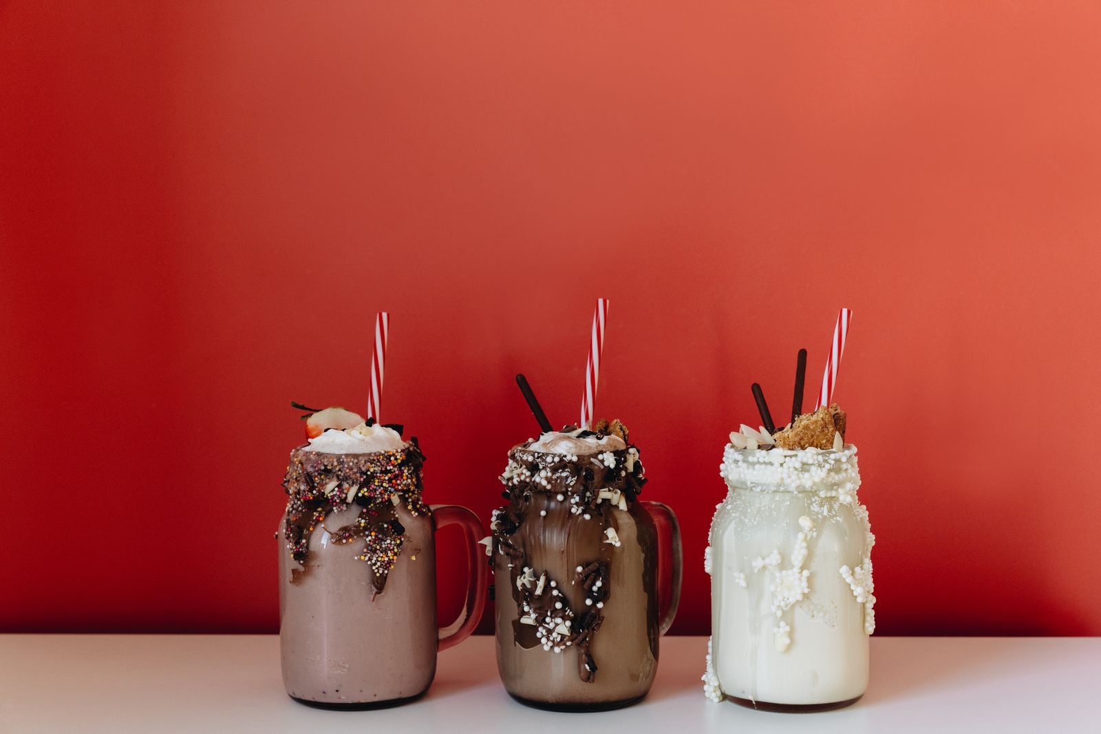 Three indulgent milkshakes in glass jars with sprinkles and chocolate against a bold red backdrop.